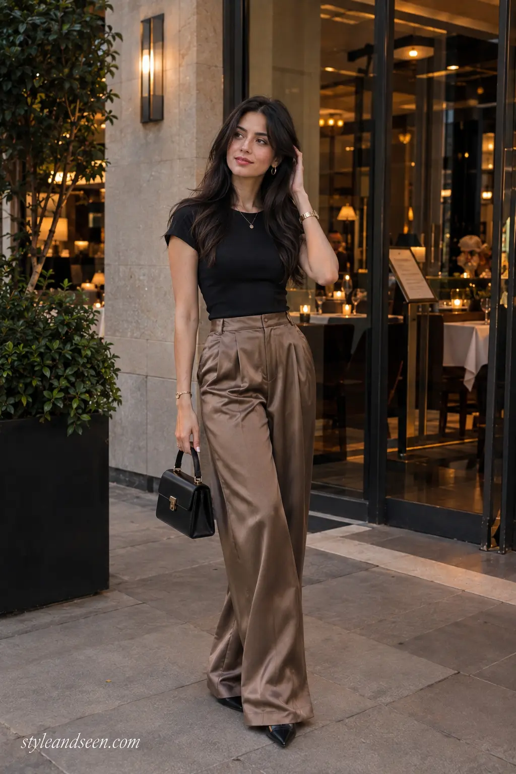 Young woman wearing a black fitted top with satin wide-leg trousers, pointed shoes, and a structured handbag for an elegant evening outfit.