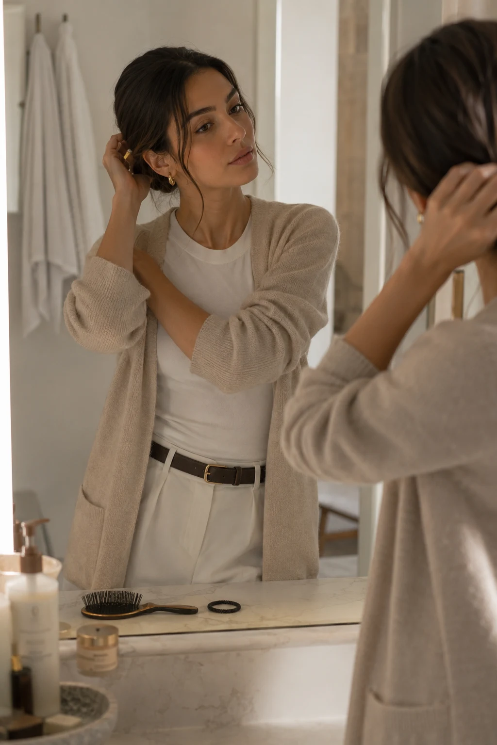 A stylish woman adjusts her polished low ponytail in front of a bathroom mirror while wearing a simple casual outfit, showing how an easy, intentional hairstyle can make a low-effort look appear polished.