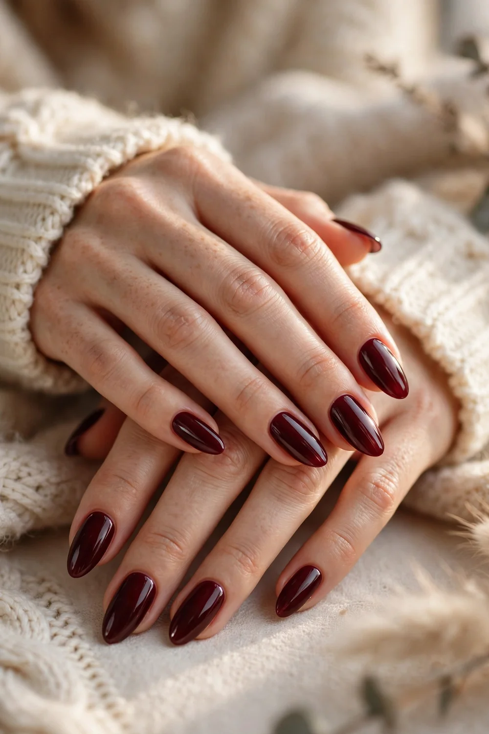 Close up of elegant hands with deep burgundy glossy almond nails and a cream knit sweater sleeve